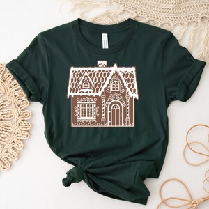 May include: A dark green t-shirt with a white gingerbread house design. The house has a brown roof, white icing details, and a brown door.