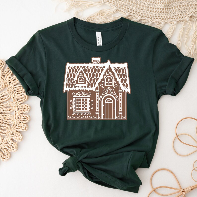 May include: A dark green t-shirt with a white gingerbread house design. The house has a brown roof, white icing details, and a brown door.