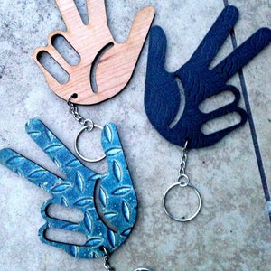 May include: Three wooden hand-shaped keychains with different designs. One is natural wood, one is black with a textured pattern, and one is blue with a diamond plate pattern.