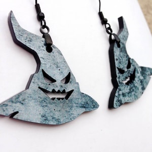 May include: A pair of silver and black earrings shaped like a ghost with a toothy grin. The earrings are made of wood and have a distressed finish.