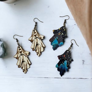 May include: Two pairs of dangle earrings with a ghost design. One pair is white with a wood grain finish and the other pair is black with a blue and green galaxy print.
