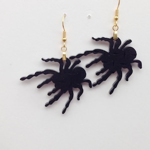 Puede incluir: Un par de pendientes de araña negros con ganchos dorados. Las arañas son detalladas y tienen un aspecto realista.