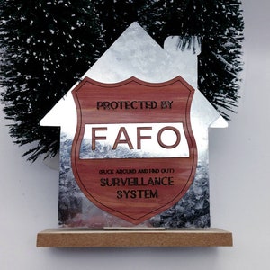 Op de afbeelding: Een zilveren huisvormig bord met een rood houten schild waarop "PROTECTED BY FAFO (F*** AROUND AND FIND OUT) SURVEILLANCE SYSTEM" staat.