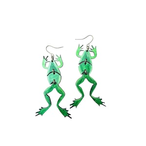 May include: A pair of green frog earrings. The frogs are made of wood and have a painted finish. The earrings are hanging from silver hooks.