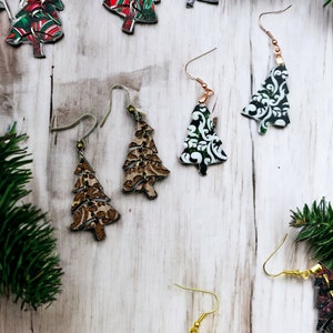 May include: A selection of Christmas tree earrings featuring different patterns and colours. The earrings are made from metal and have a hook fastening. The earrings are shown on a white wooden background.