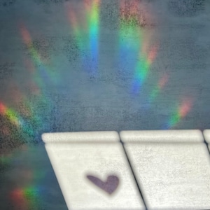 Puede incluir: Un objeto rectangular blanco con un dise&ntilde;o en forma de coraz&oacute;n p&uacute;rpura en primer plano. El fondo presenta una superficie gris texturizada con un efecto de luz de arco&iacute;ris. El efecto arco&iacute;ris tiene bandas de rojo, naranja, amarillo, verde, azul y morado.
