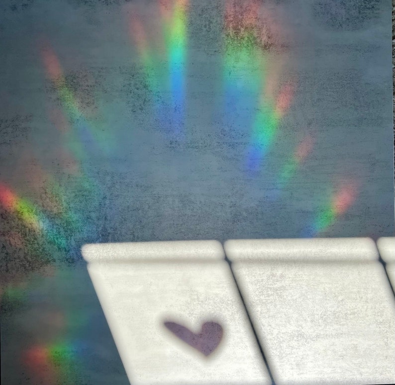 Puede incluir: Un objeto rectangular blanco con un dise&ntilde;o en forma de coraz&oacute;n p&uacute;rpura en primer plano. El fondo presenta una superficie gris texturizada con un efecto de luz de arco&iacute;ris. El efecto arco&iacute;ris tiene bandas de rojo, naranja, amarillo, verde, azul y morado.