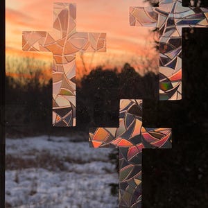 May include: Three decorative crosses with stained glass-like patterns are affixed to a window. The crosses are in a cross shape, with a colorful mosaic design. The background shows a sunset over a snowy landscape.