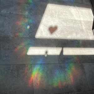 Puede incluir: Fotograf&iacute;a de un suelo embaldosado gris con un espectro de luz de arco&iacute;ris y una sombra en forma de coraz&oacute;n. La luz crea una exhibici&oacute;n colorida en el suelo, con la sombra del coraz&oacute;n agregando un elemento &uacute;nico.