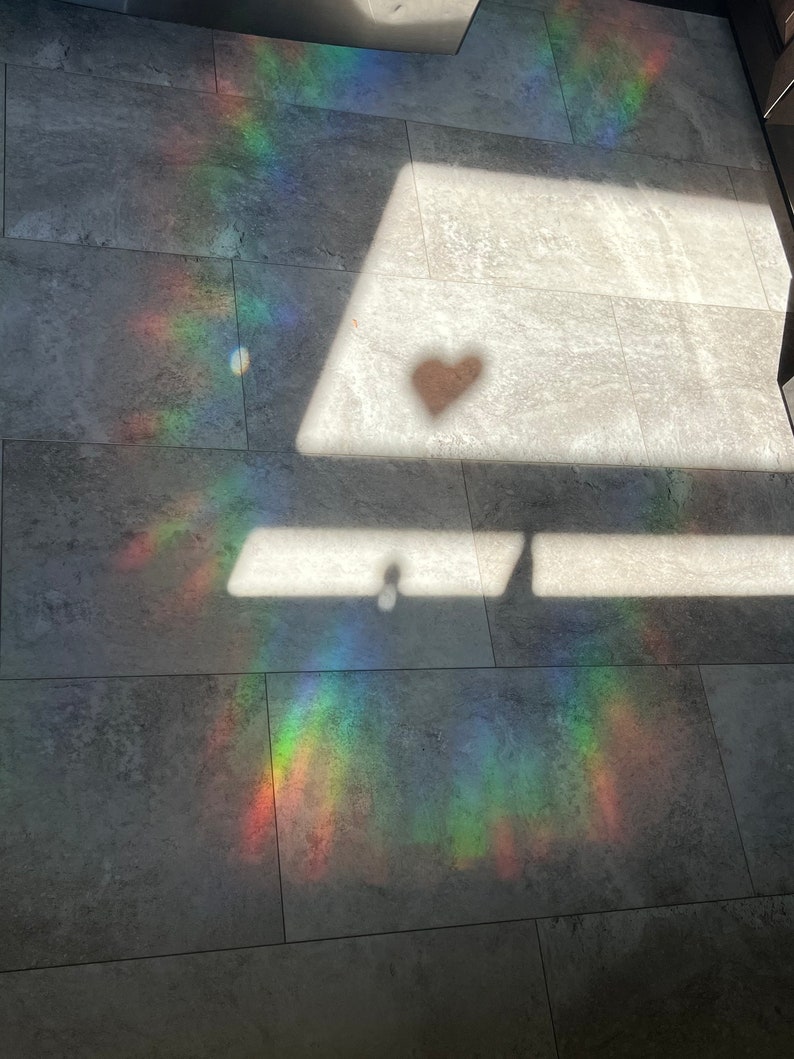 Puede incluir: Fotograf&iacute;a de un suelo embaldosado gris con un espectro de luz de arco&iacute;ris y una sombra en forma de coraz&oacute;n. La luz crea una exhibici&oacute;n colorida en el suelo, con la sombra del coraz&oacute;n agregando un elemento &uacute;nico.