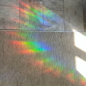 Puede incluir: Un vibrante espectro de luz de arco&iacute;ris se proyecta sobre un suelo de baldosas grises y una alfombra beige. La colorida exhibici&oacute;n incluye bandas de rojo, naranja, amarillo, verde, azul y violeta, creando un efecto visual impactante.