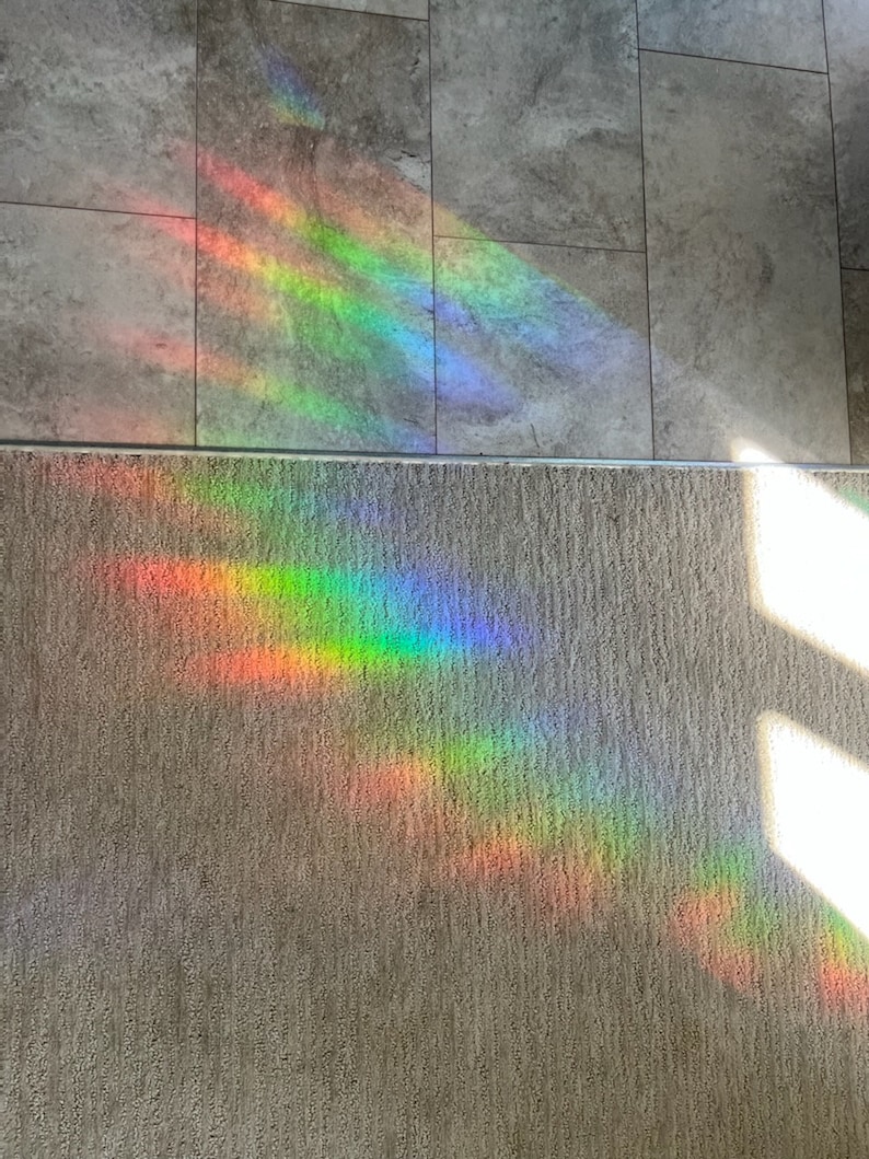 Puede incluir: Un vibrante espectro de luz de arco&iacute;ris se proyecta sobre un suelo de baldosas grises y una alfombra beige. La colorida exhibici&oacute;n incluye bandas de rojo, naranja, amarillo, verde, azul y violeta, creando un efecto visual impactante.