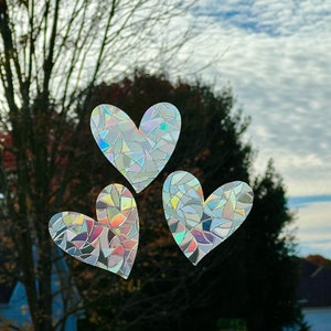 Puede incluir: Tres pegatinas en forma de coraz&oacute;n con un dise&ntilde;o de mosaico est&aacute;n adheridas a una ventana. Las pegatinas tienen un efecto iridiscente de arco&iacute;ris, que refleja varios colores. El fondo muestra un cielo nublado y &aacute;rboles.