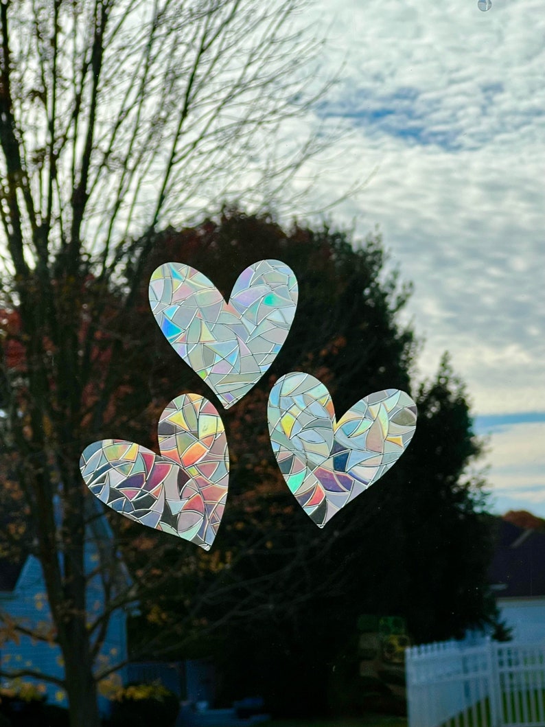 Puede incluir: Tres pegatinas en forma de coraz&oacute;n con un dise&ntilde;o de mosaico est&aacute;n adheridas a una ventana. Las pegatinas tienen un efecto iridiscente de arco&iacute;ris, que refleja varios colores. El fondo muestra un cielo nublado y &aacute;rboles.
