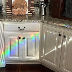 Puede incluir: Armarios de cocina blancos con pomos negros y encimera de granito. Un espectro de luz de arco&iacute;ris se proyecta sobre los armarios. Se ven una olla de acero inoxidable y una tabla de cortar de madera.