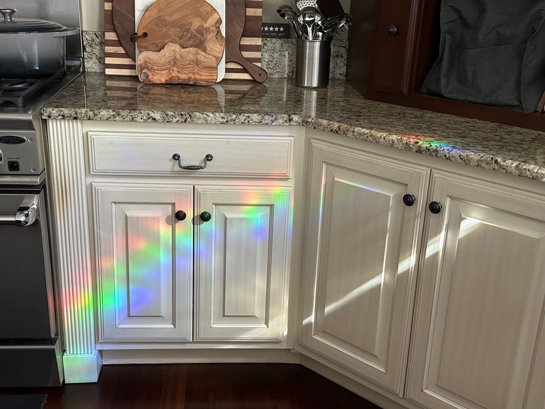 Puede incluir: Armarios de cocina blancos con pomos negros y encimera de granito. Un espectro de luz de arco&iacute;ris se proyecta sobre los armarios. Se ven una olla de acero inoxidable y una tabla de cortar de madera.