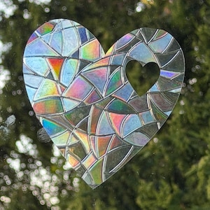 Puede incluir: Una pegatina para ventana en forma de corazón con efecto vidriera. La pegatina presenta un patrón de mosaico de colores iridiscentes, incluyendo azul, verde, rojo y amarillo. Una forma de corazón más pequeña está recortada del centro. El fondo es una vista borrosa de follaje verde.