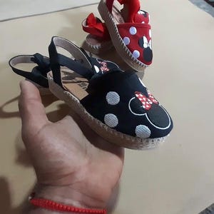 Puede incluir: Dos pares de sandalias de esparto con dise&ntilde;o de Minnie Mouse. Un par es negro con lunares blancos y el otro es rojo con lunares blancos.