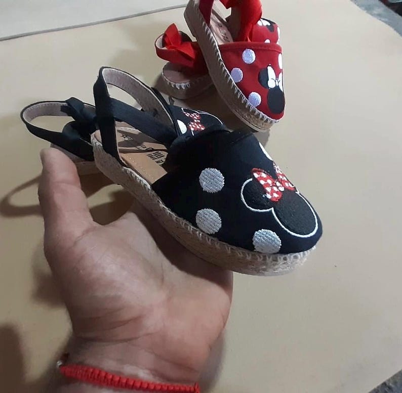 Puede incluir: Dos pares de sandalias de esparto con dise&ntilde;o de Minnie Mouse. Un par es negro con lunares blancos y el otro es rojo con lunares blancos.
