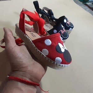 Puede incluir: Dos pares de sandalias de esparto con partes superiores de lona roja y negra. El par rojo tiene lunares blancos y un lazo rojo con orejas negras. El par negro tiene lunares blancos y un lazo negro con orejas rojas.