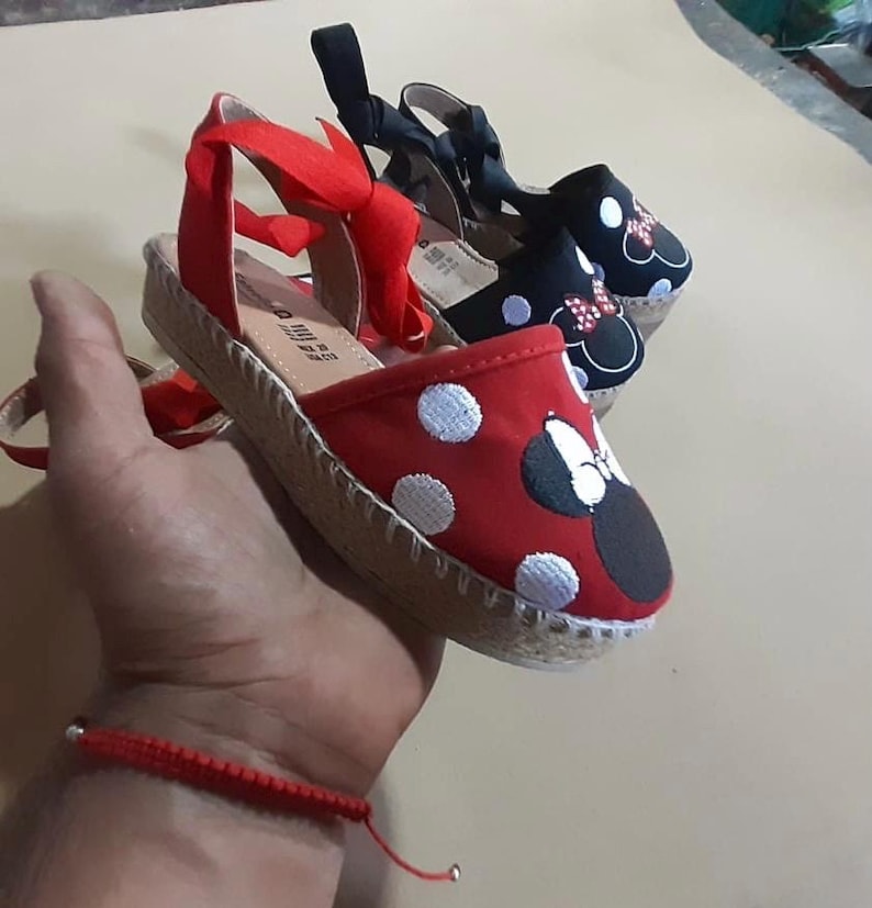 Puede incluir: Dos pares de sandalias de esparto con partes superiores de lona roja y negra. El par rojo tiene lunares blancos y un lazo rojo con orejas negras. El par negro tiene lunares blancos y un lazo negro con orejas rojas.