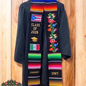 May include: Black graduation gown with a colorful serape-style sash. The sash features the Puerto Rican and Mexican flags, the text "CLASS OF 2026", and the name "JIMENEZ". The sash is adorned with floral embroidery and the letters "UNT".