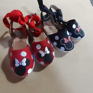Puede incluir: Dos pares de zapatos de esparto de lona roja y negra con lunares blancos y un dise&ntilde;o bordado de Minnie Mouse en blanco y negro. Los zapatos tienen cordones de cinta roja y negra.