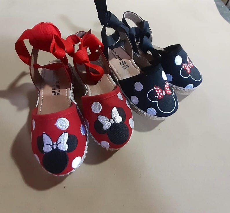 Puede incluir: Dos pares de zapatos de esparto de lona roja y negra con lunares blancos y un dise&ntilde;o bordado de Minnie Mouse en blanco y negro. Los zapatos tienen cordones de cinta roja y negra.