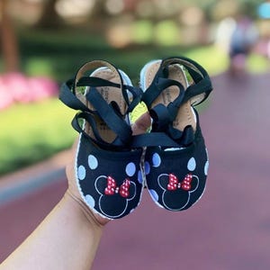Puede incluir: Zapatillas de esparto de lunares negros y blancos con lazos rojos y un dise&ntilde;o de Minnie Mouse. Las zapatillas tienen cordones de cinta negra.