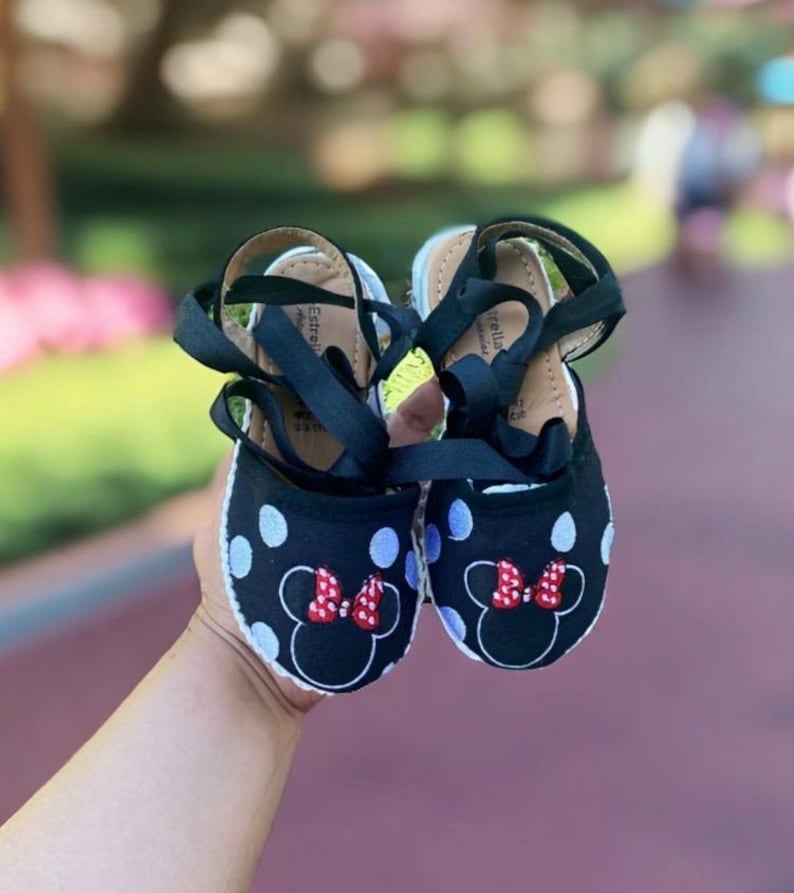 Puede incluir: Zapatillas de esparto de lunares negros y blancos con lazos rojos y un dise&ntilde;o de Minnie Mouse. Las zapatillas tienen cordones de cinta negra.