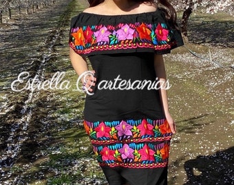 May include: A black off-the-shoulder dress with colorful floral embroidery. The dress has a ruffled neckline and a fitted bodice. The embroidery features flowers in shades of pink, red, orange, yellow, and blue. The dress is photographed in front of a row of trees with white blossoms.