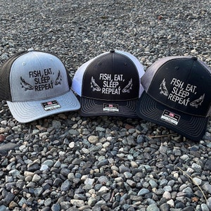 Puede incluir: Tres gorras de camionero negras y grises con una malla blanca en la parte posterior. Las gorras tienen un diseño bordado en negro que dice "Fish, Eat, Sleep, Repeat" con un gráfico de pescado.