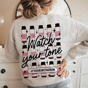 May include: Grey sweatshirt with a graphic design of pink and black bottles and the text "Watch your tone" and "@yourinstagram".