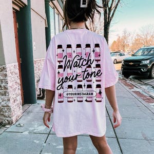 Puede incluir: Camiseta rosa de gran tama&ntilde;o con un gr&aacute;fico de botellas de colores y el texto "Watch your tone" y "@yourinstagram".