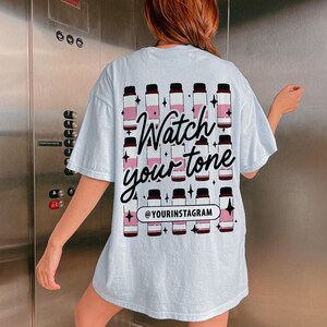 Puede incluir: Camiseta azul claro con un gr&aacute;fico de botellas rosas y negras y el texto "Watch your tone" y "@yourinstagram".