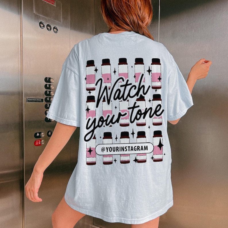 Puede incluir: Camiseta azul claro con un gr&aacute;fico de botellas rosas y negras y el texto "Watch your tone" y "@yourinstagram".