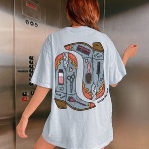 Puede incluir: Camiseta azul claro con un gr&aacute;fico de dos botas de vaquero, cada una con herramientas de peluquer&iacute;a. Las botas est&aacute;n delineadas en marr&oacute;n y naranja, con el texto "CUSTOM TEXT HERE" en la bota derecha.
