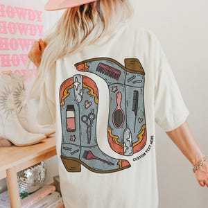 Puede incluir: Camiseta color crema con una ilustraci&oacute;n estilizada de dos botas de vaquero. Las botas son de color azul claro con detalles en naranja y marr&oacute;n, y contienen herramientas de peluquer&iacute;a. El texto "CUSTOM TEXT HERE" est&aacute; impreso en la bota derecha.