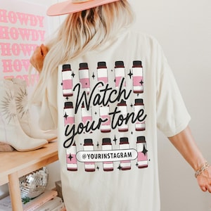 Puede incluir: Una camiseta blanca con un dise&ntilde;o gr&aacute;fico rosa y marr&oacute;n de botellas y el texto "Watch your tone" y "@yourinstagram".