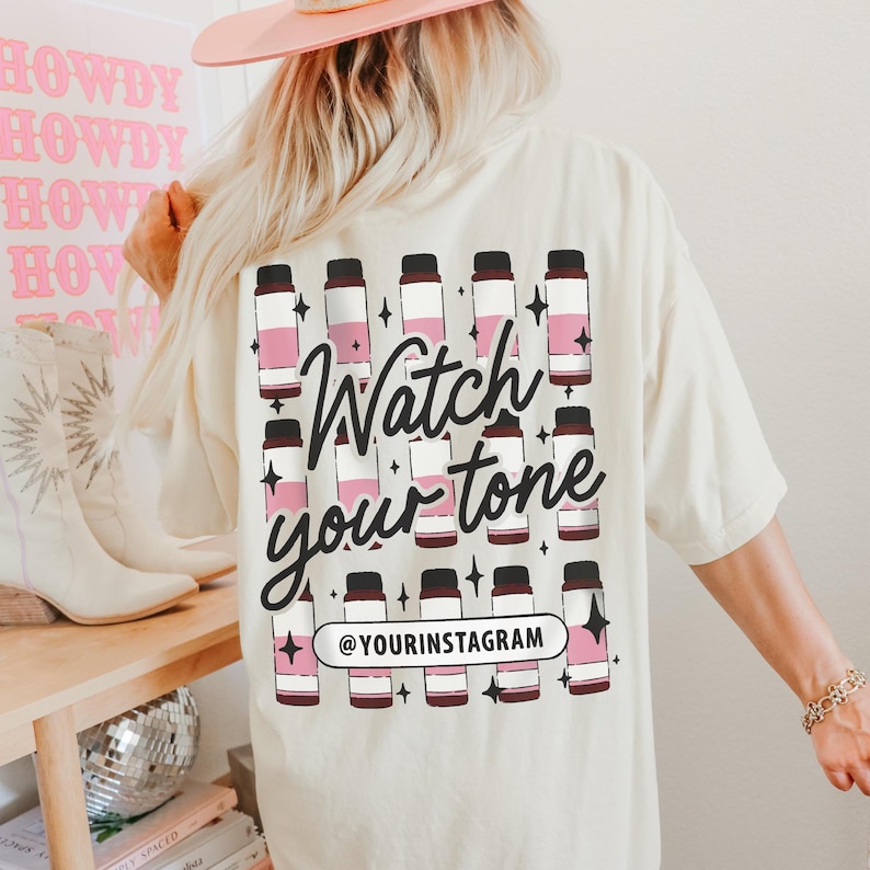Puede incluir: Una camiseta blanca con un dise&ntilde;o gr&aacute;fico rosa y marr&oacute;n de botellas y el texto "Watch your tone" y "@yourinstagram".