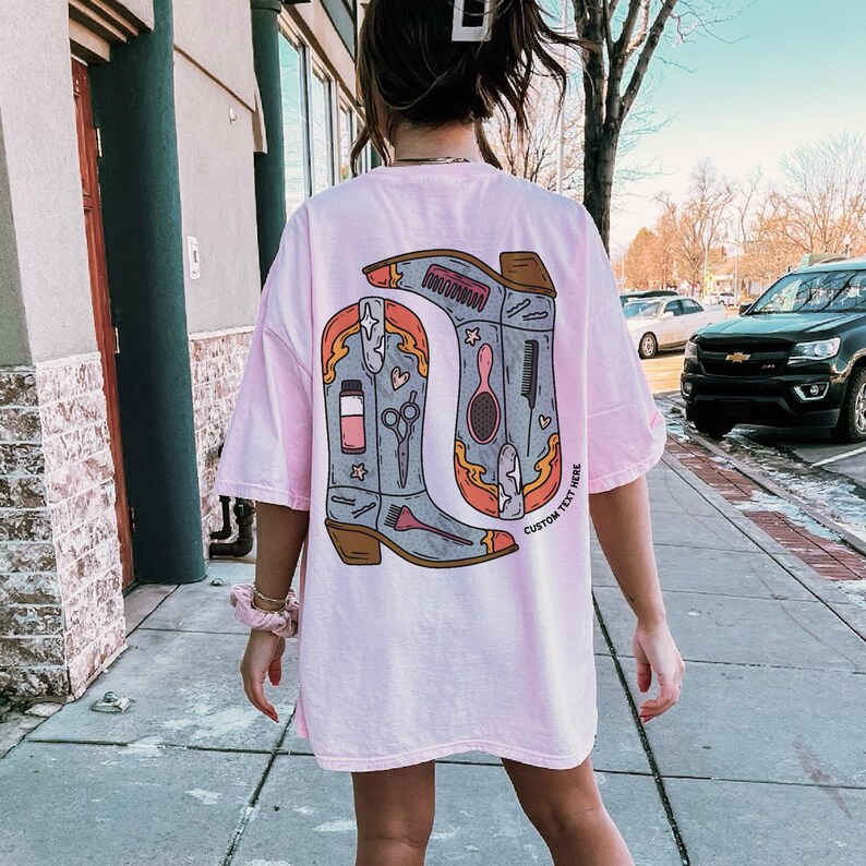 Puede incluir: Camiseta rosa claro de gran tama&ntilde;o con un gr&aacute;fico de dos botas de vaquero con herramientas de sal&oacute;n. Las botas est&aacute;n delineadas en naranja y marr&oacute;n, con el texto "CUSTOM TEXT HERE" en la bota derecha.