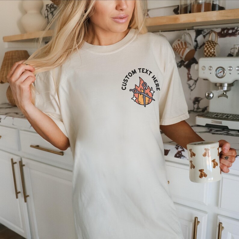Puede incluir: Camiseta color crema con un gr&aacute;fico en la parte delantera que dice "CUSTOM TEXT HERE". El gr&aacute;fico presenta tijeras, llamas y estrellas. Una taza blanca con ilustraciones de osos marrones se sostiene en la mano.