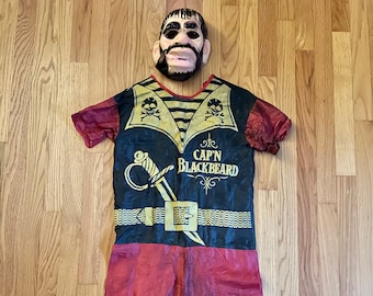 May include: A vintage Captain Blackbeard costume for kids, featuring a red and black shirt with a gold pirate sword and skull and crossbones design, red pants, and a plastic mask with a beard.