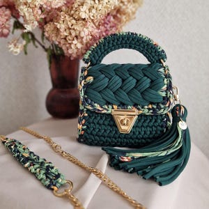 May include: A dark green crocheted handbag featuring a braided flap, a gold-tone metal turn-lock closure, and a matching fabric tassel. The bag includes a detachable gold chain strap with a crocheted shoulder pad in green, black, and yellow floral print.