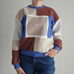 Könnte beinhalten: Ein farblich abgesetztes Sweatshirt mit Patchwork-Design in Blau-, Braun- und Beigetönen. In der Mitte der Brust befindet sich eine quadratische Grafik mit dem weißen Text "bye.". Das Kleidungsstück hat einen gerippten blauen Rundhalsausschnitt.