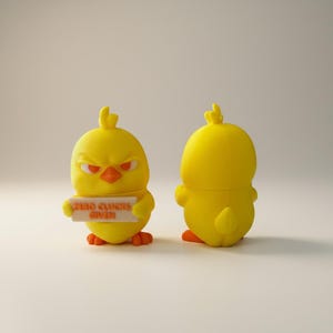 May include: Two yellow chick figurines. One chick has an angry expression, red eyes, and an orange beak. It holds a sign that reads "ZERO CLUCKS GIVEN." The second chick is facing away. Both have orange feet.
