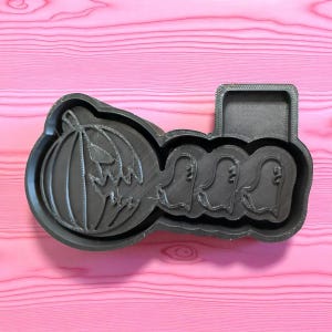 May include: A black plastic Halloween-themed cookie cutter. The design includes a carved pumpkin, three ghosts, and a rectangular area. The cutter is on a pink wooden surface.