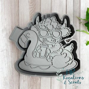May include: A gray plastic cookie cutter in the shape of a cartoon cow wearing sunglasses and floating on a flamingo pool float. The cow is holding a drink with a straw. The cutter has a wavy border and the text "HR Kreations & Scents".