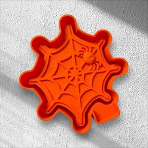 May include: Bright orange Halloween-themed cookie cutter in the shape of a spider web with a small spider. The cutter has a scalloped edge and is made of plastic. Perfect for making spooky treats.