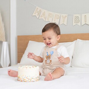 May include: A beige romper featuring an embroidered brown teddy bear wearing a blue party hat and holding a blue balloon with the number one. The romper is worn with a white short-sleeved shirt. A white cake with colorful sprinkles sits nearby.